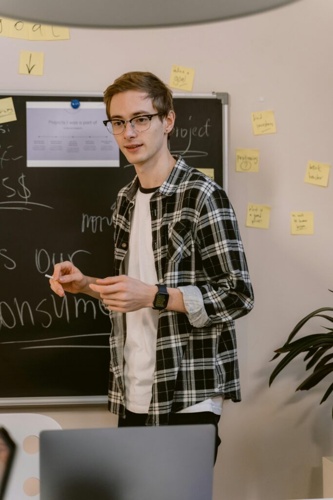 Young man in casual attire with eyeglasses, presenting in front of a chalkboard covered with sticky notes.